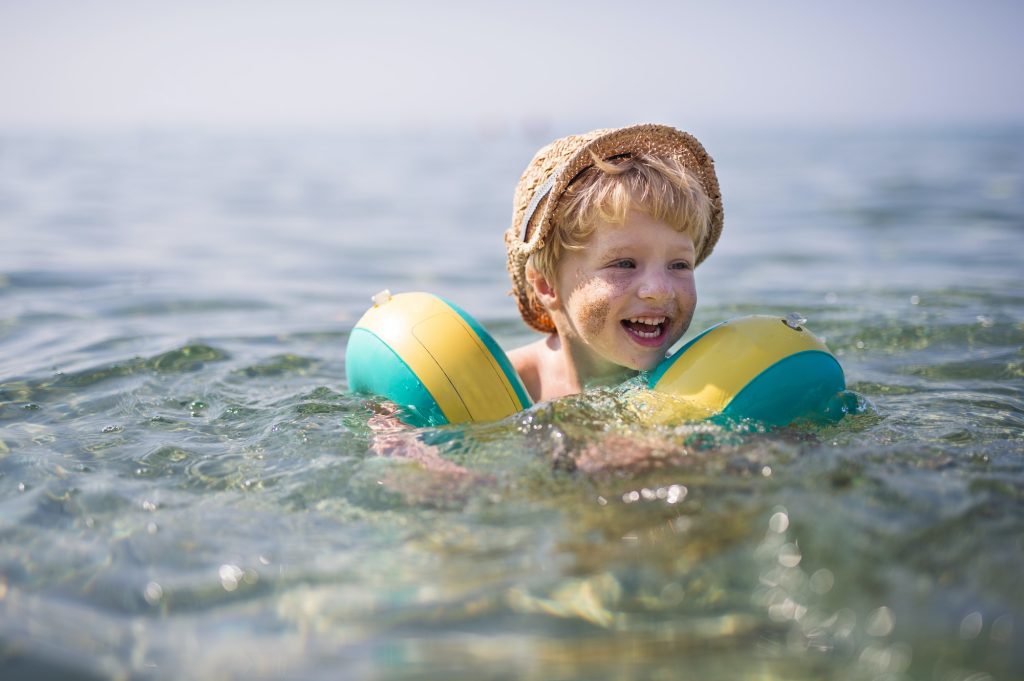 A small toddler boy with armbands swimming in water on summer holiday.