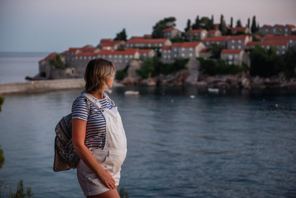 Adult pregnant woman in vest, white overalls stands by the sea. The concept of motherhood in travel