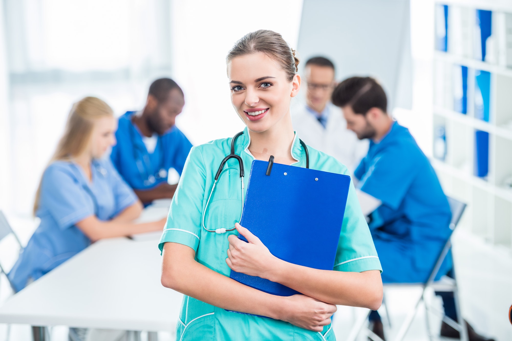 attractive young nurse holding clipboard with colleagues sitting at table on background