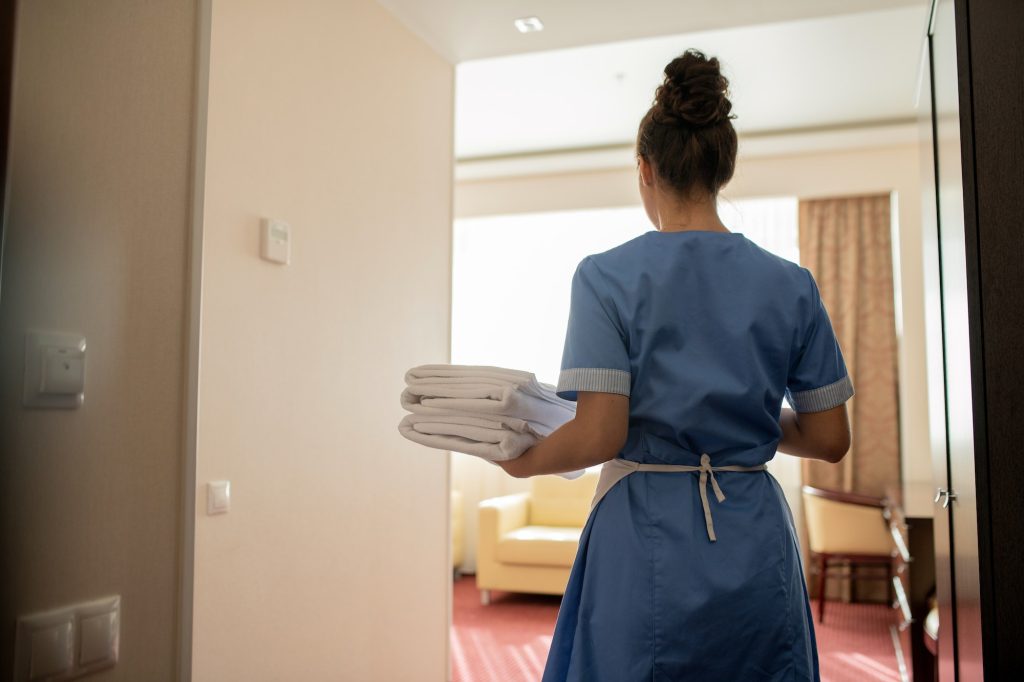 Back view of young chamber maid in uniform carrying stack of clean white towels