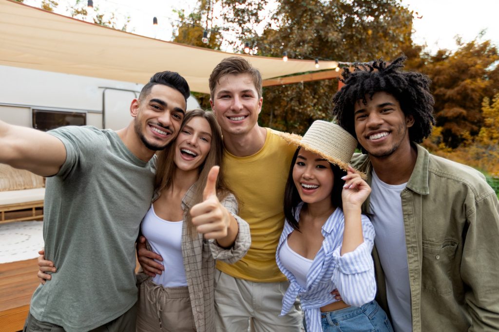 Diverse millennials taking selfie together near camper van, smiling at camera, spending time