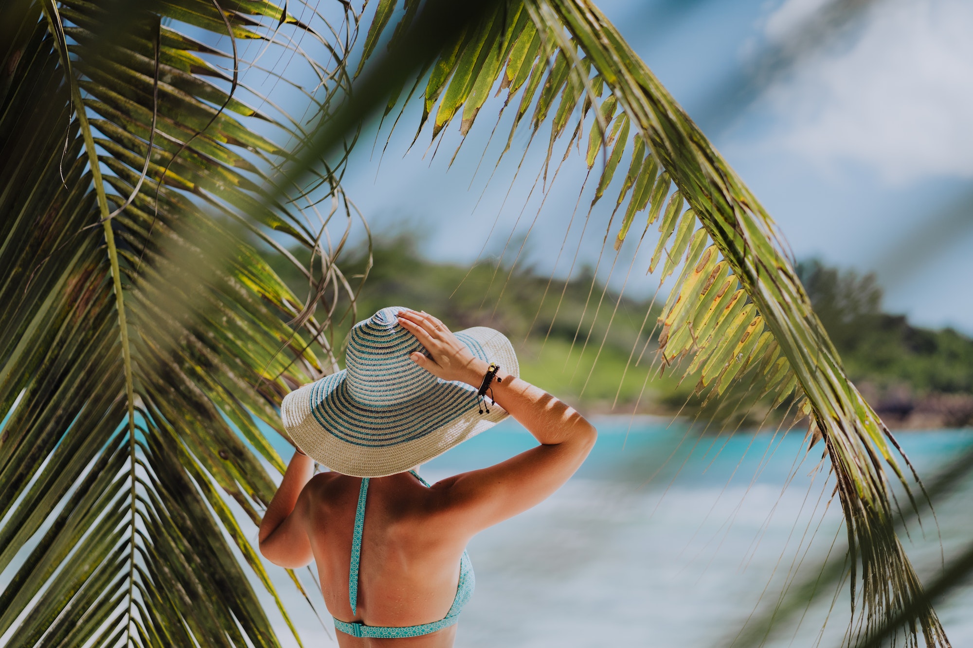 Woman on the beach in the palm trees shadow wearing blue hat. Luxury paradise recreation vacation